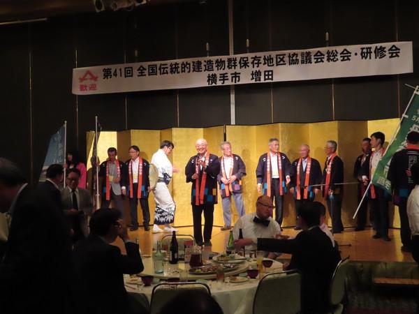 舞台で法被を着た男性達が並んで、市長が代表でお話ししている写真